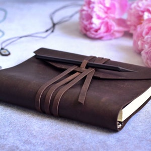 May include: A dark brown leather journal with a pencil secured by leather straps. The journal is closed, with a flap and a pencil tucked in. Pink peonies and a necklace are in the background.