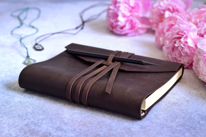 May include: A dark brown leather journal with a pencil secured by leather straps. The journal is closed, with a flap and a pencil tucked in. Pink peonies and a necklace are in the background.