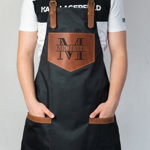 May include: A black apron with brown leather accents and adjustable straps. The apron has a large pocket on the front with a personalized leather patch that says "MICHAEL" with a monogrammed "M".