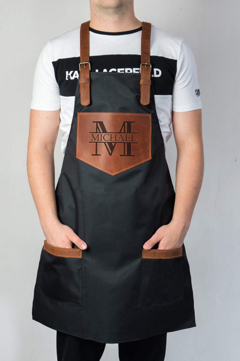 May include: A black apron with brown leather accents and adjustable straps. The apron has a large pocket on the front with a personalized leather patch that says "MICHAEL" with a monogrammed "M".