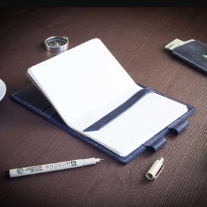 Op de afbeelding: Een blauw leren notitieboekje met een wit papieren inlegvel en een pen op een houten tafel. Het notitieboekje heeft een leren sluiting met een riem en een pennenhouder.