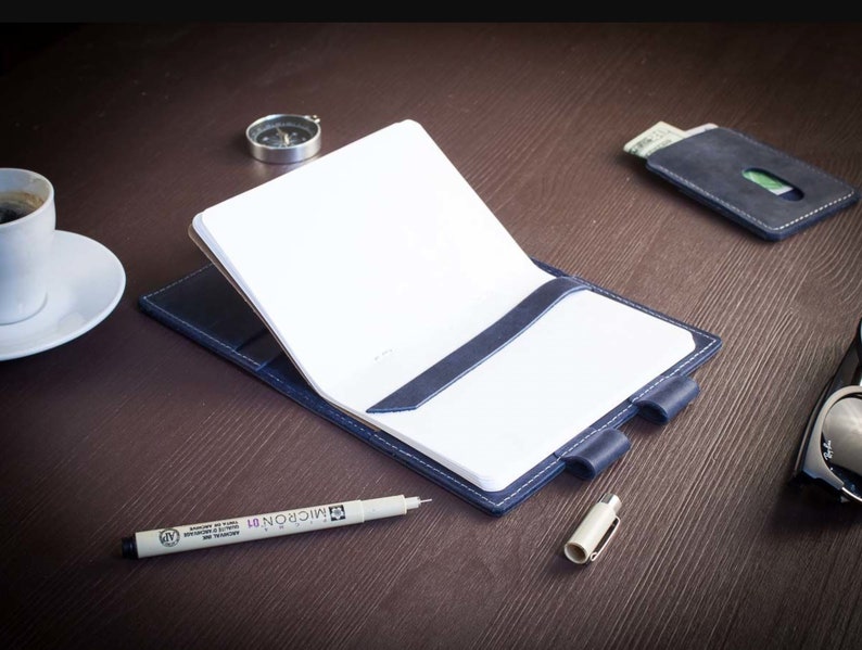 Op de afbeelding: Een blauw leren notitieboekje met een wit papieren inlegvel en een pen op een houten tafel. Het notitieboekje heeft een leren sluiting met een riem en een pennenhouder.