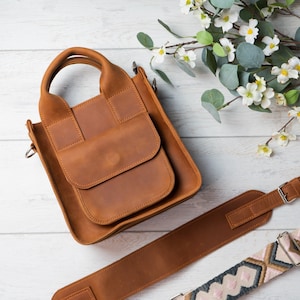 May include: A brown leather tote bag with a top handle and a long adjustable strap. The bag has a flap closure and a front pocket.