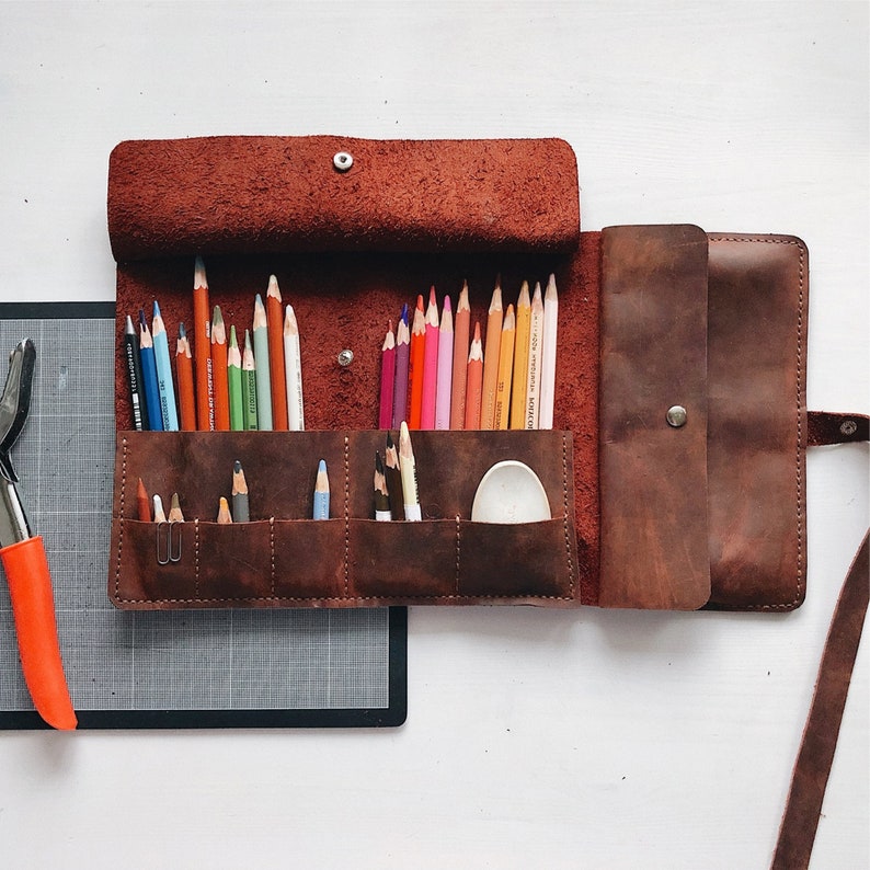 May include: A brown leather pencil case with a flap closure and a zippered pocket. The case is open and filled with colored pencils. There is a small white plastic container inside the case.