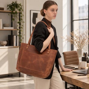 Puede incluir: Un bolso tote grande y rectangular de cuero color marr&oacute;n co&ntilde;ac, con dos asas largas sujetas por remaches met&aacute;licos dorados y un bolsillo exterior ancho y prominente en la parte frontal, con una textura de cuero natural, lisa y mate.