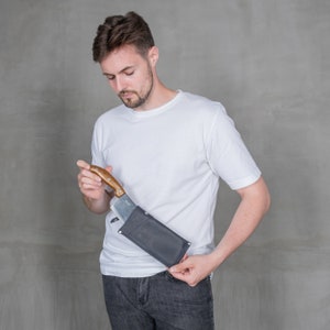 May include: A person wearing a white t-shirt and blue jeans is holding a knife in a black leather sheath.