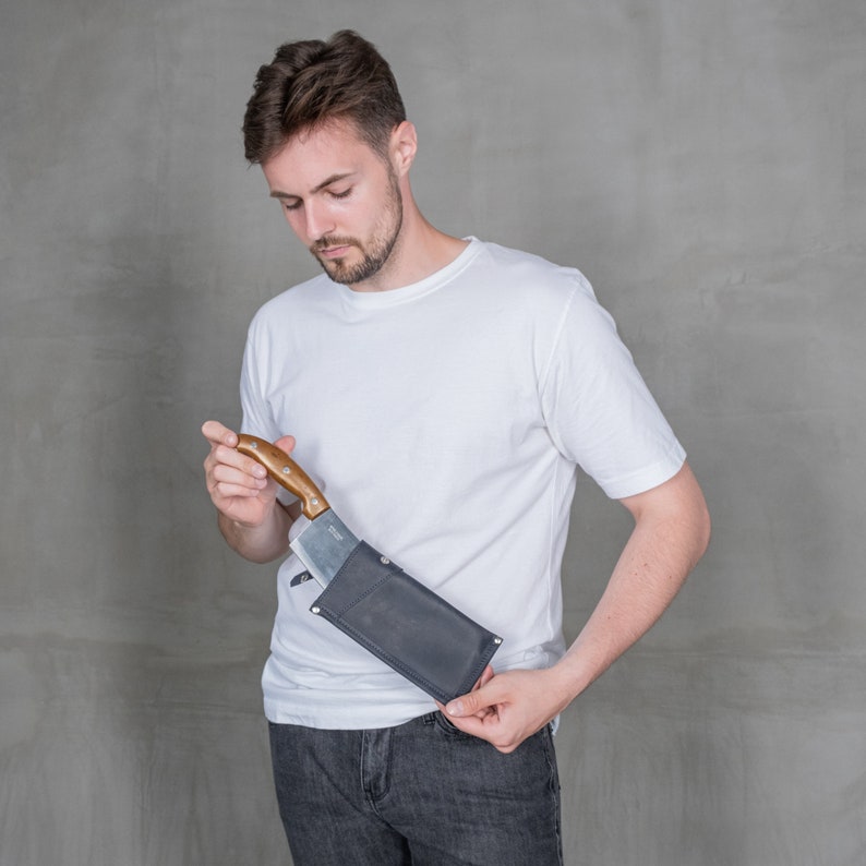 May include: A person wearing a white t-shirt and blue jeans is holding a knife in a black leather sheath.