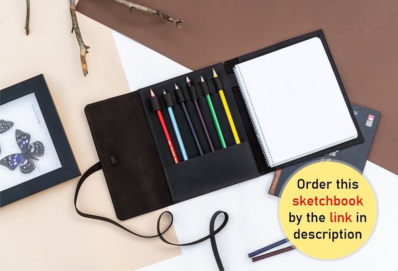 May include: A brown leather sketchbook with a built-in pencil holder. The sketchbook has a spiral binding and is closed. The pencil holder has seven coloured pencils: red, blue, light blue, black, dark green, light green, and yellow. The text "Order this sketchbook by the link in description" is visible in the image.