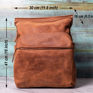 May include: A brown leather backpack with two zippered compartments and a top flap. The backpack measures 16 inches tall, 11.8 inches wide, and 6.3 inches deep.