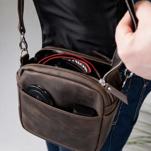 May include: A brown leather crossbody bag with a zippered main compartment and an open front pocket. The bag contains a black camera lens with a red strap and a black lens cap. It features silver-toned metal hardware and a thin adjustable shoulder strap.
