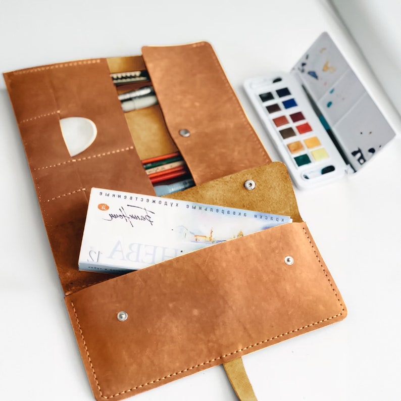 May include: A brown leather roll-up case with multiple compartments for holding art supplies. The case is open and a small book with the text "ФЕВА" is visible inside. A set of watercolour paints is also visible in the background.