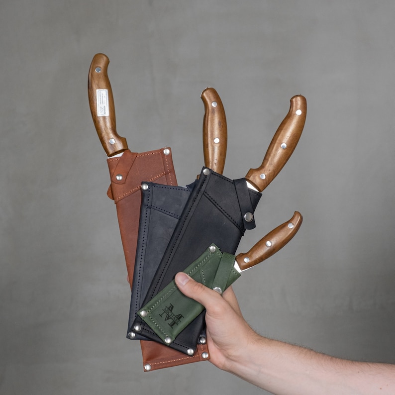 May include: Three leather sheaths with wooden-handled knives. The sheaths are brown, black, and green. The knives have a wooden handle and a metal blade.