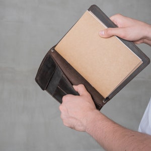 May include: A brown leather notebook cover with a flap that opens to reveal a blank notebook. The notebook is held in a person's hand.