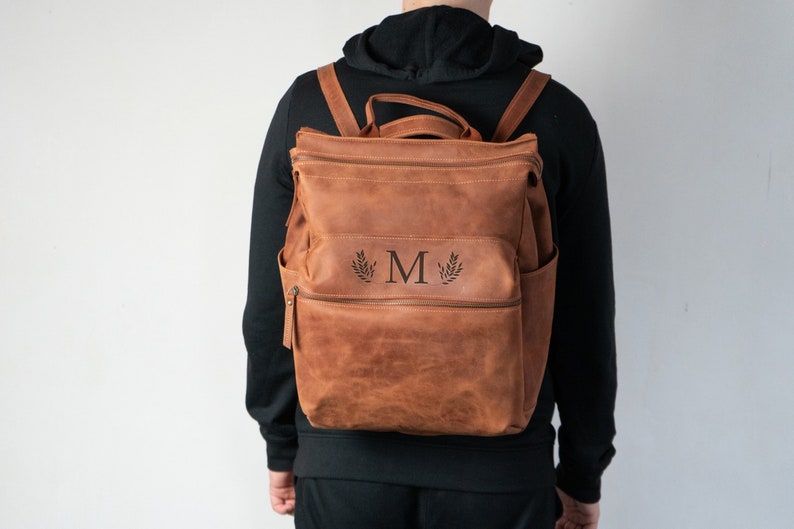 May include: A brown leather backpack with a monogrammed initial "M" and a leafy design. The backpack has a zippered pocket on the front and adjustable straps.