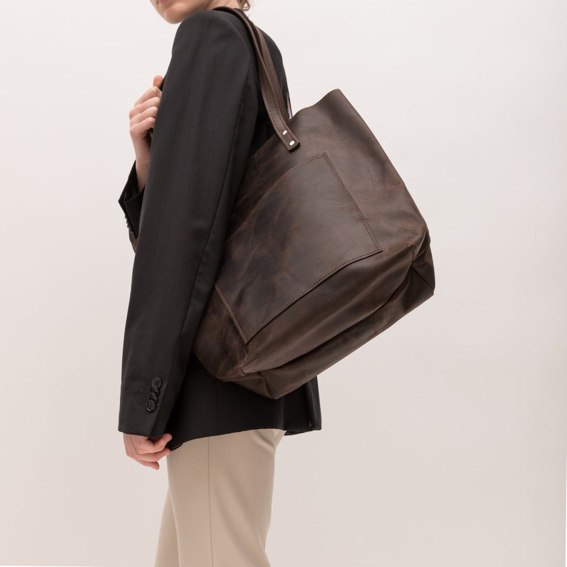 Puede incluir: Un bolso tote grande de cuero marr&oacute;n oscuro con un compartimento principal espacioso, un bolsillo frontal rectangular y dos correas de hombro resistentes sujetas con remaches met&aacute;licos visibles. El cuero tiene un acabado mate y desgastado.