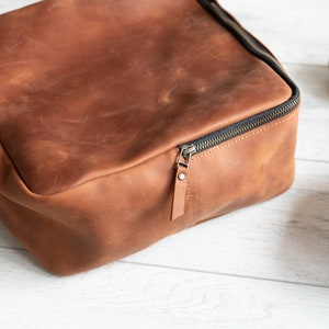 May include: A brown leather lunch bag with a zipper closure. The bag is sitting on a white wooden surface.