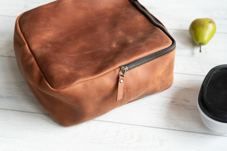 May include: A brown leather lunch bag with a zipper closure. The bag is sitting on a white wooden surface.