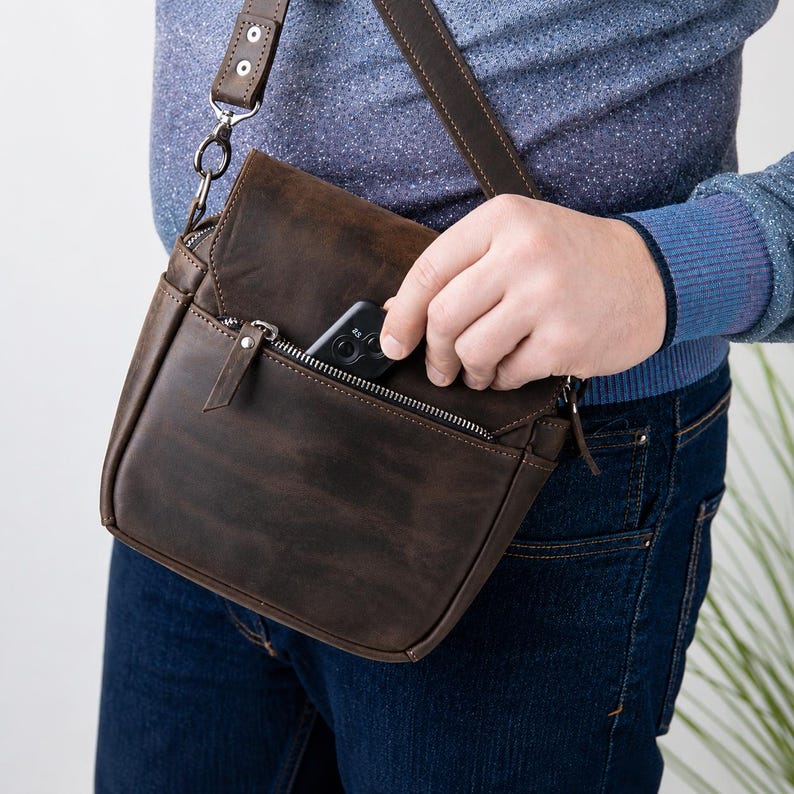 May include: A dark brown leather crossbody bag featuring a front zippered pocket, a fold-over flap with a magnetic closure, and an adjustable shoulder strap. The bag has visible stitching details and silver-toned metal hardware accents.