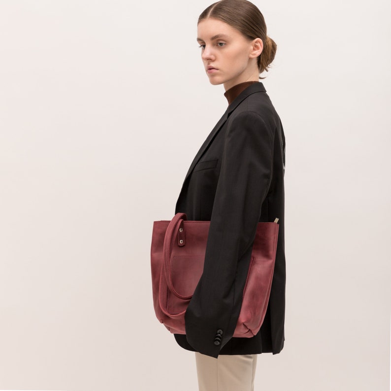 Puede incluir: Un bolso tote de cuero rojo oscuro con silueta rectangular, dos correas de hombro sujetas con remaches de metal plateado y un bolsillo exterior visible. El bolso tiene un acabado mate y liso, mostrado desde una vista de perfil.