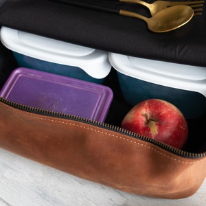 May include: A brown leather zippered pouch with a black lining. The pouch is open and contains two clear plastic containers, a purple container, and a red apple.