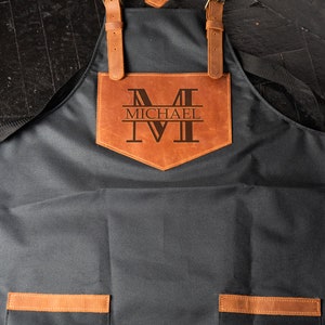 May include: A black canvas apron with brown leather straps and a large brown leather pocket on the front. The pocket has the letter "M" and the name "MICHAEL" engraved on it.