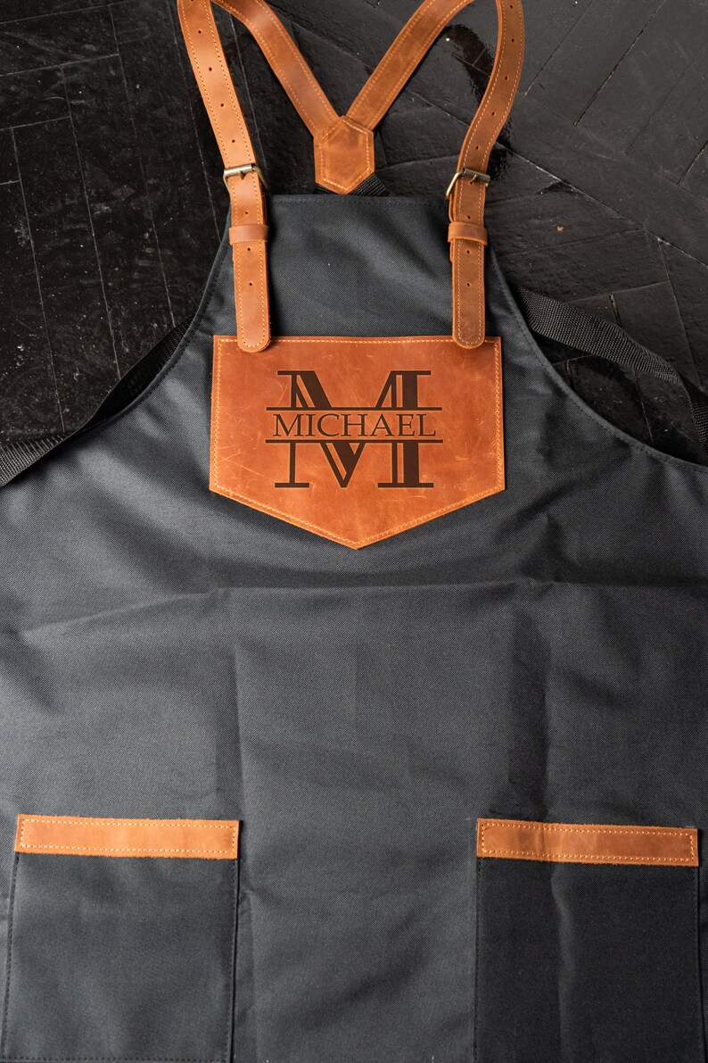 May include: A black canvas apron with brown leather straps and a large brown leather pocket on the front. The pocket has the letter "M" and the name "MICHAEL" engraved on it.