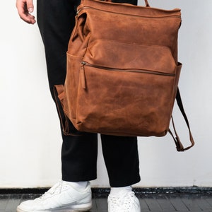 May include: A brown leather backpack with a flap top and a zippered pocket. The backpack has a top handle and adjustable straps.