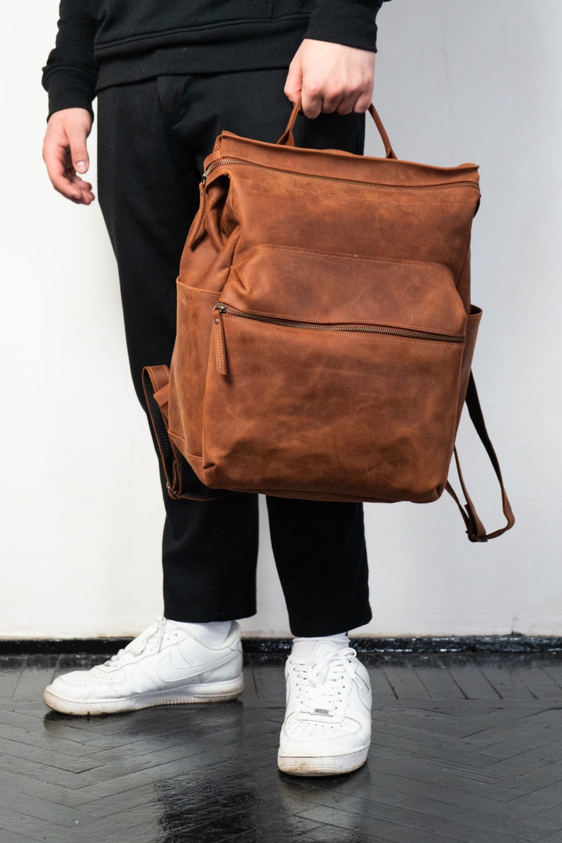 May include: A brown leather backpack with a flap top and a zippered pocket. The backpack has a top handle and adjustable straps.