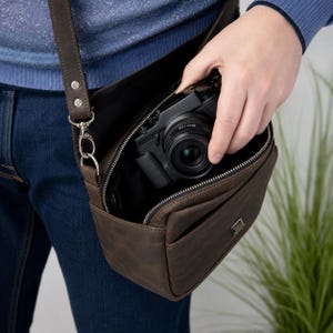 May include: A dark brown leather camera bag with a zippered main compartment and an adjustable shoulder strap attached with silver metal hardware. The bag features a front pocket and is shown containing a black digital camera with a lens.