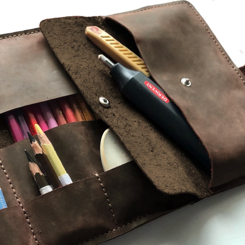 May include: A brown leather pencil case with multiple slots for pencils and other art supplies. The case is open and shows a wooden pencil sharpener, a black pencil sharpener with the brand name "DERWENT" printed on it, and a set of coloured pencils.