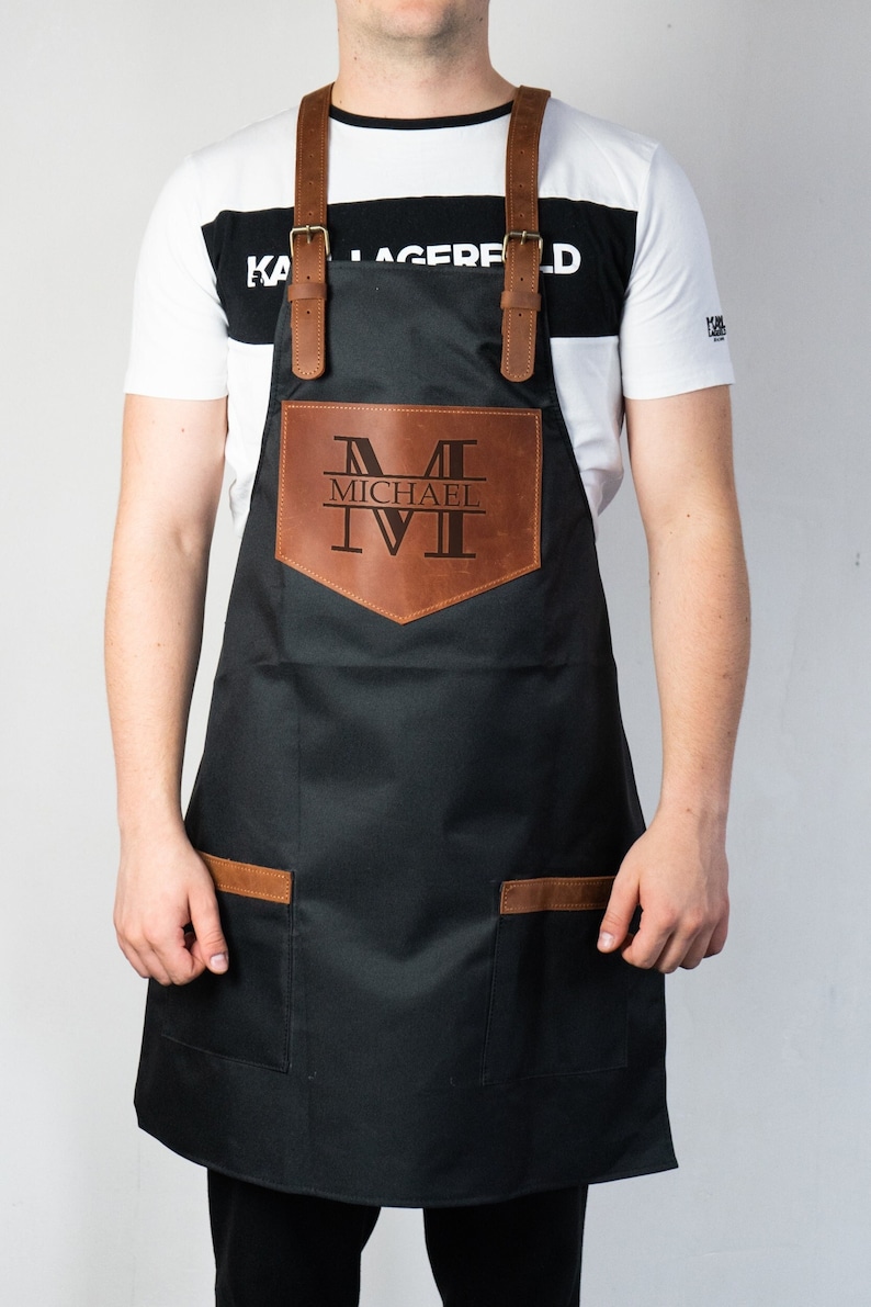 May include: A black canvas apron with brown leather straps and pockets. The apron has a large brown leather patch on the front with the name "MICHAEL" and the letter "M" in Roman numerals.