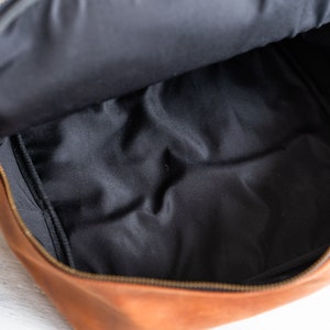 May include: A brown leather bag with a black fabric lining. The bag is open and the lining is visible. The bag has a zipper closure.