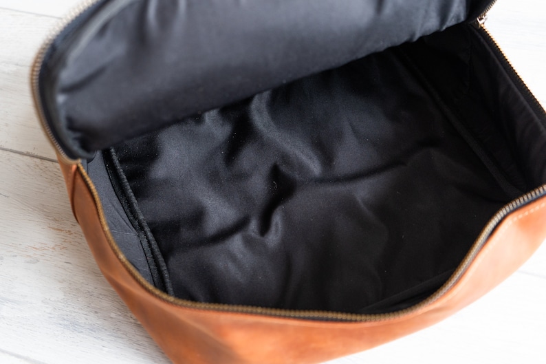 May include: A brown leather bag with a black fabric lining. The bag is open and the lining is visible. The bag has a zipper closure.