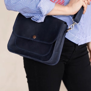 Pu&ograve; includere: Una borsa a tracolla in pelle blu scuro con chiusura a lembo e una piccola tasca sul davanti. La borsa ha un bottone marrone e una fibbia argentata sulla tracolla.