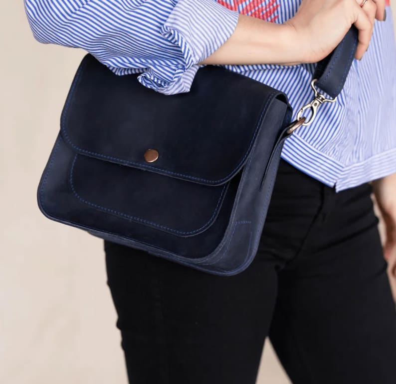 Pu&ograve; includere: Una borsa a tracolla in pelle blu scuro con chiusura a lembo e una piccola tasca sul davanti. La borsa ha un bottone marrone e una fibbia argentata sulla tracolla.