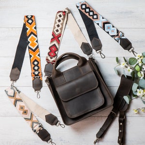 May include: A brown leather tote bag with a top flap closure and a front pocket. The bag has three different patterned straps, one with orange and white stripes, one with blue and white stripes, and one with red and white stripes.