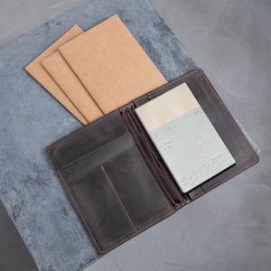 Könnte beinhalten: Ein braunes Leder-Notizbuchcover mit Reißverschluss und mehreren Taschen. Das Cover ist geöffnet und zeigt ein leeres Notizbuch mit einem Rastermuster und dem Text "MD NOTE" in japanischer Sprache.