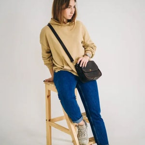 Pu&ograve; includere: Una donna indossa una felpa beige, jeans blu e scarpe da ginnastica bianche e siede su uno sgabello di legno. Porta una borsa a tracolla in pelle marrone.