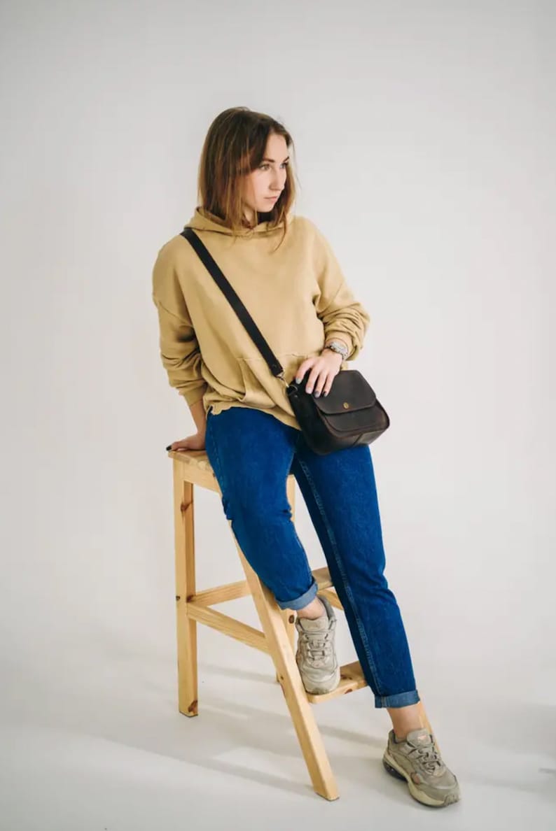 Pu&ograve; includere: Una donna indossa una felpa beige, jeans blu e scarpe da ginnastica bianche e siede su uno sgabello di legno. Porta una borsa a tracolla in pelle marrone.