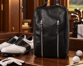 May include: A black leather shoe bag with a top handle and two vertical silver zippered pockets on the front. The bag is positioned next to a pair of white and black golf shoes, a white golf glove, two golf balls, and a scorecard.