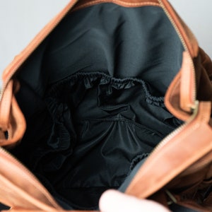 May include: A brown leather diaper bag with a black interior and a zipper closure. The bag has a flap with a buckle closure and a strap.