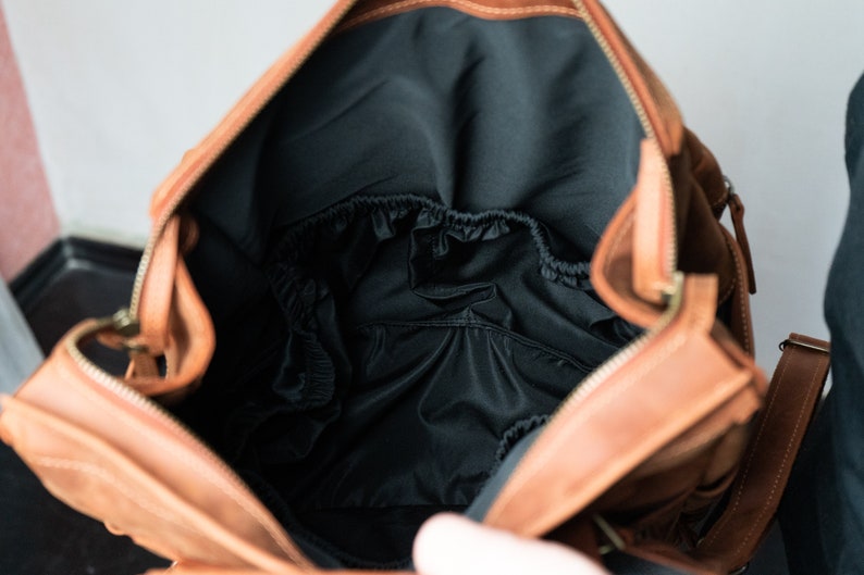 May include: A brown leather diaper bag with a black interior and a zipper closure. The bag has a flap with a buckle closure and a strap.