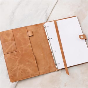 Op de afbeelding: Een beige lederen ordner met een metalen mechanisme met vier ringen, een verticale riemsluiting met drukknoop en een linker binnenpaneel met twee gestapelde vakken en een penlus, gevuld met vellen wit papier.