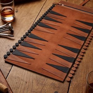 May include: A roll-up backgammon board made of brown leather featuring alternating black and brown triangular points. The set includes a row of black circular checkers and a row of brown circular checkers arranged along the sides of the board.