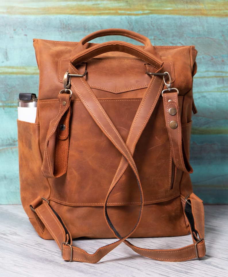 May include: A brown leather backpack with adjustable straps and a zippered pocket. The backpack has a top handle and a side pocket.