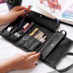 May include: Black leather roll-up case with multiple compartments for holding art supplies, such as pencils, pens, and markers. The case is open and the compartments are filled with various art supplies.