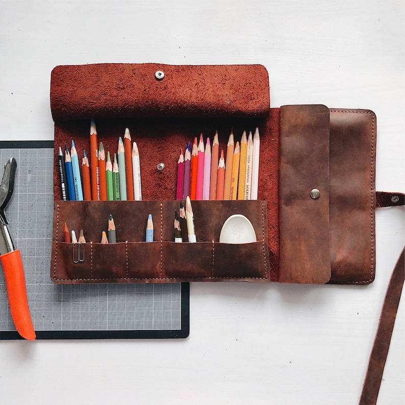 May include: A brown leather pencil case with a flap closure and a zipped pocket. The case is open and filled with coloured pencils. There is a small white plastic container inside the case.