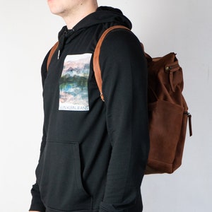 May include: A person wearing a black hoodie with a graphic print of a landscape and the text "Calvin Klein Jeans". The person is wearing a brown leather backpack.