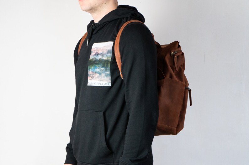 May include: A person wearing a black hoodie with a graphic print of a landscape and the text "Calvin Klein Jeans". The person is wearing a brown leather backpack.