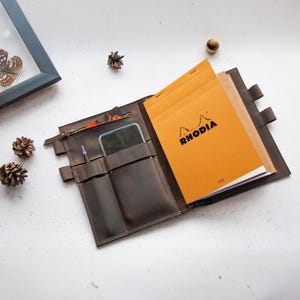 Puede incluir: Una cubierta de cuaderno de cuero marrón con cierre de cremallera, varios bolsillos y un portaplumas. La cubierta está abierta para revelar un cuaderno Rhodia amarillo con el nombre de la marca impreso en la parte delantera.